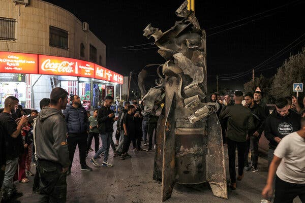 A crowd stands outside shops and around a large projectile in open space.