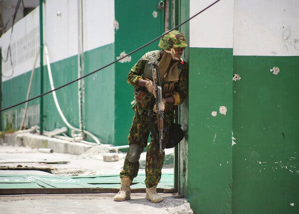 A man dressed in military clothing and with a gun leans against a pocked green and white building.