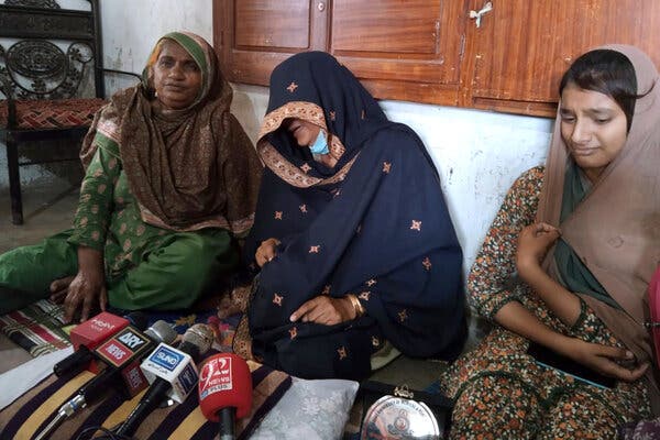 Three women in Pakistan sit on the ground, in front of a row of microphones. 