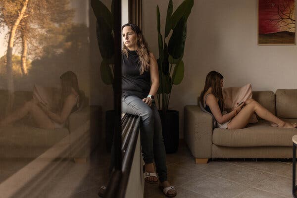 A woman in a black top gazes out the window while her daughter sits and reads on a couch nearby. 