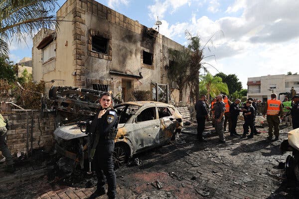 A woman in uniform standing near a charred car and a damaged building. Other officials, some in orange vests, are nearby.