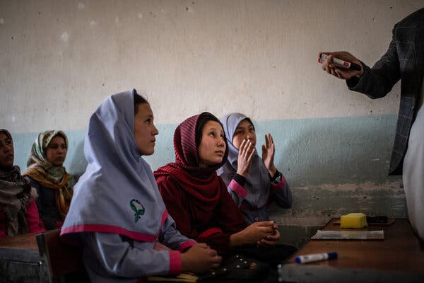 Women wearing head coverings sitting at tables and looking up as someone speaks to them.