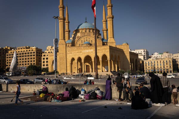 Several people sit or stand with their belongings in a parking lot across the street from a mosque.