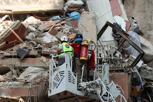 Two people wearing helmets survey a tangle of concrete rubble, wires and household belongings at the site of a strike.