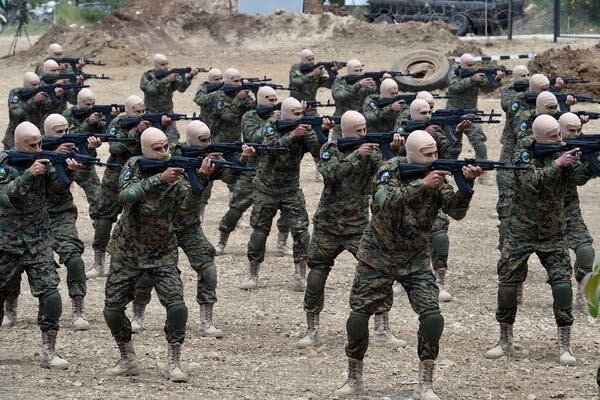 Men in camouflage and wearing khaki-colored masks aiming weapons.