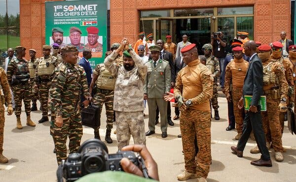 Men in military uniforms, some wearing red caps, gathered in front of a building. A man in the center of the group is raising his arms and clasping his hands. 