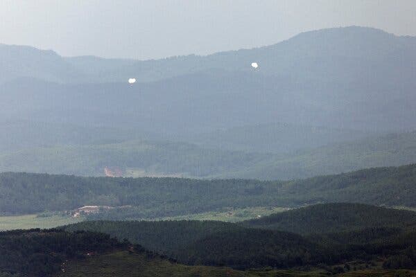 Two white balloons float in the distance over a green, hilly landscape.