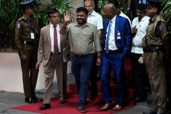 A man in blue pants and a taupe shirt waves as he walks on a red carpet, flanked and followed by other men.