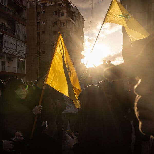 People holding a yellow flag on the street, with large buildings and a sunset in the background.