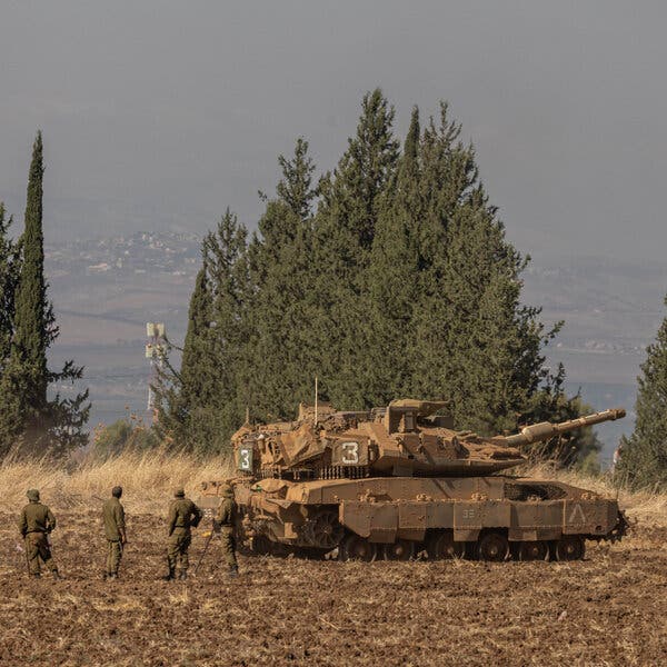 Soldiers next to a tank in a field, with a line of trees behind and a hillside seen through haze beyond that.