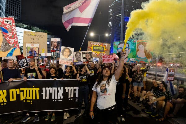 A crowd of people hold up posters and a banner Saturday in Tel Aviv calling for a hostage deal. One person raises an arm, releasing yellow smoke above the crowd. 