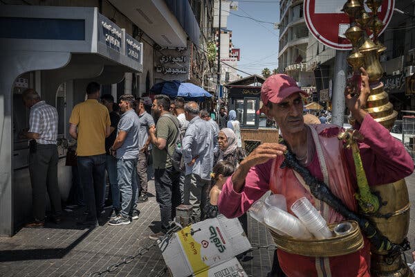 People lining up on a sidewalk by a bank machine in a busy city area. In the foreground stands a man with a large brass jug and stacks of plastic cups.