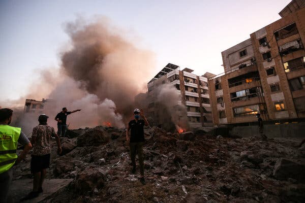 Thick smoke rises near multistory buildings near rubble as four people stand nearby.