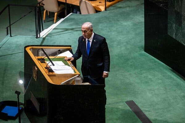 Prime Minister Benjamin Netanyahu stands on a green carpet behind a lectern, which has papers placed atop it.