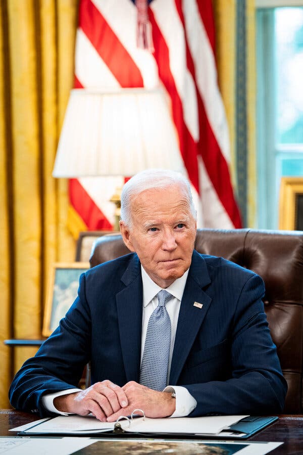 President Biden, wearing a blue suit and blue tie, sits at a desk. An open binder is in front of him, and an American flag is in the background.