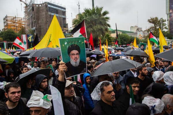 A group of people holding yellow flags, umbrellas and a photo of an older man gather outside.