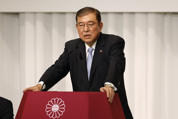 A man in a suit stands at a lectern, leaning on it with both hands.