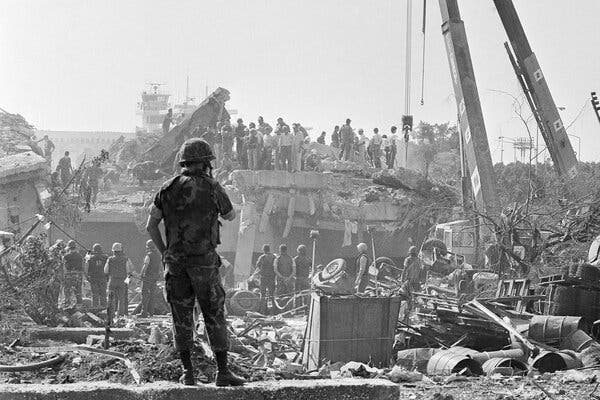 A person wearing camouflage clothing and helmet looks to the distance at a dilapidated building and people standing on the rubble.