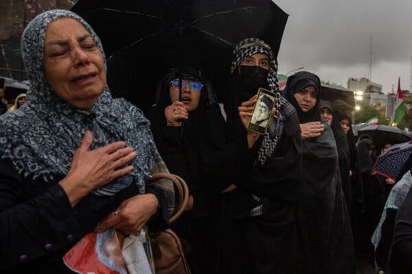 Women in a crowd mourning. One woman holds an umbrella and another a phone displaying a person's face.