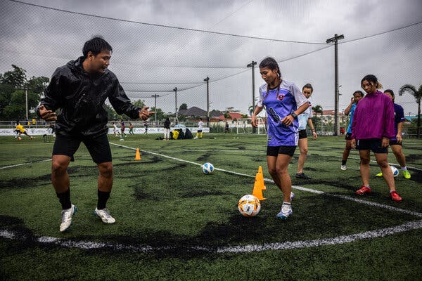 A woman in shorts and a soccer jersey prepares to kick a soccer ball that a man nearby has apparently just kicked to her. Several other women look on.