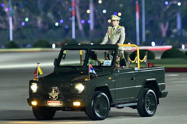 A man wearing a military uniform and a lot of medals stands up in an open-top military vehicle.