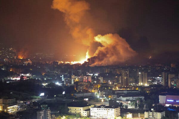 A distant view of a city at night, with a bright explosion and billowing smoke on the horizon.