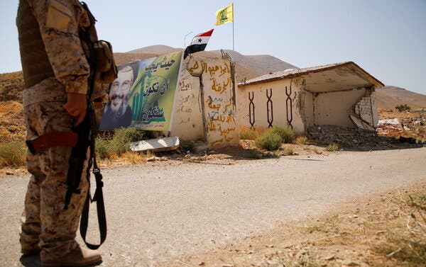 A uniformed soldier holding an automatic weapon stands near a destroyed building on a dusty road. 