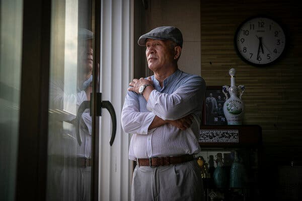 A man wearing a cap stands with arms folded in a residential setting. A clock and vase is visible behind him. 