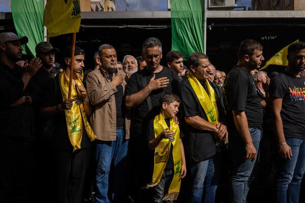 A group of somber adults and a small boy, some with their hands on their hearts, some wearing yellow shawls.