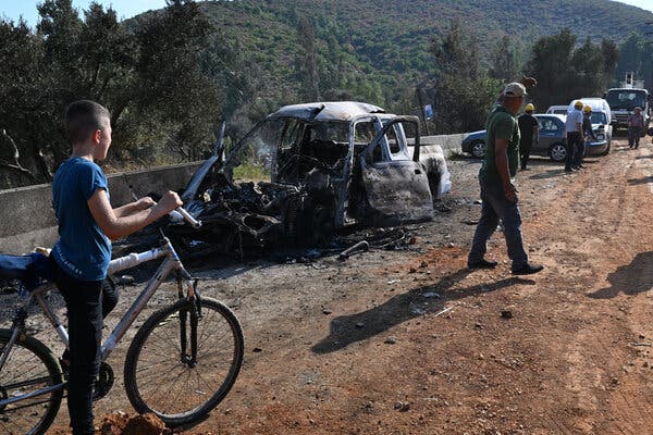 A boy on a bike looks at a burned-out car as men in hats inspect the site.