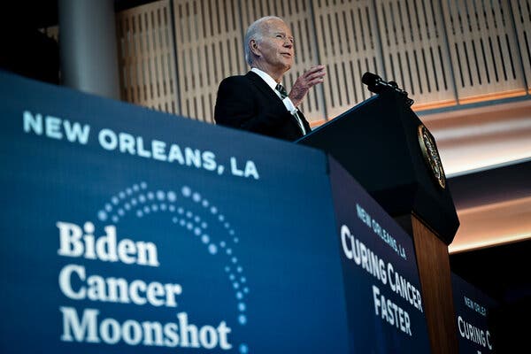 President Biden, wearing a dark suit, stands at a lectern. In the foreground is a sign with the words “Biden Cancer Moonshot.”