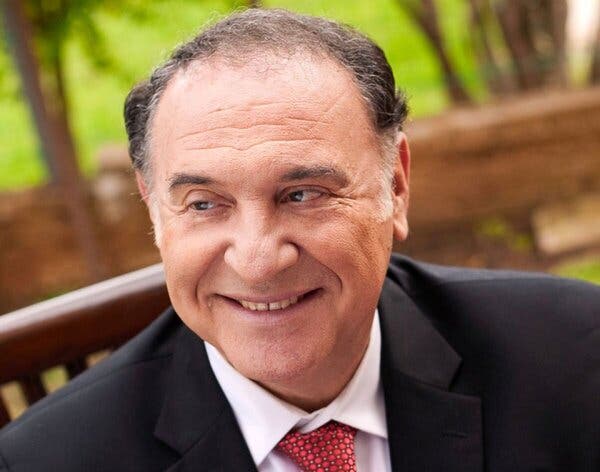 Naftali Herstik, a man with thinning gray hair wearing a blue blazer and a red tie, sits on a bench outdoors and smiles.