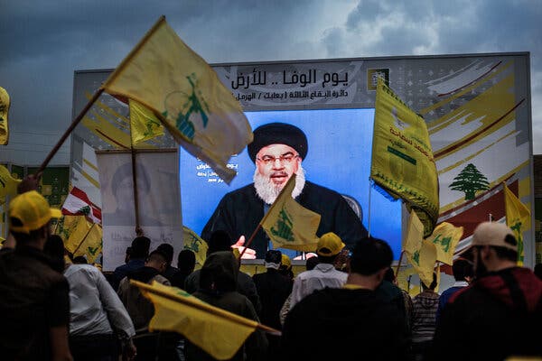 A large screen, showing Hassan Nasrallah, seen through a crowd waving flags.