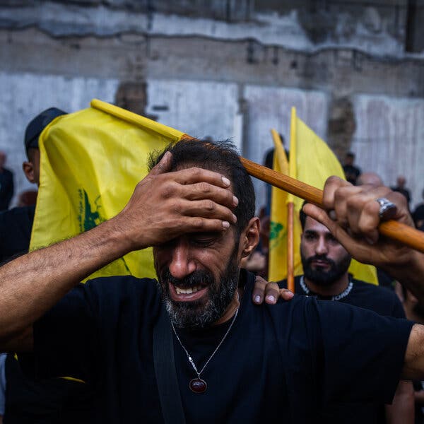 A man carrying a yellow flag stands at the front of a crowd. He holds his palm to his forehead and looks distraught.