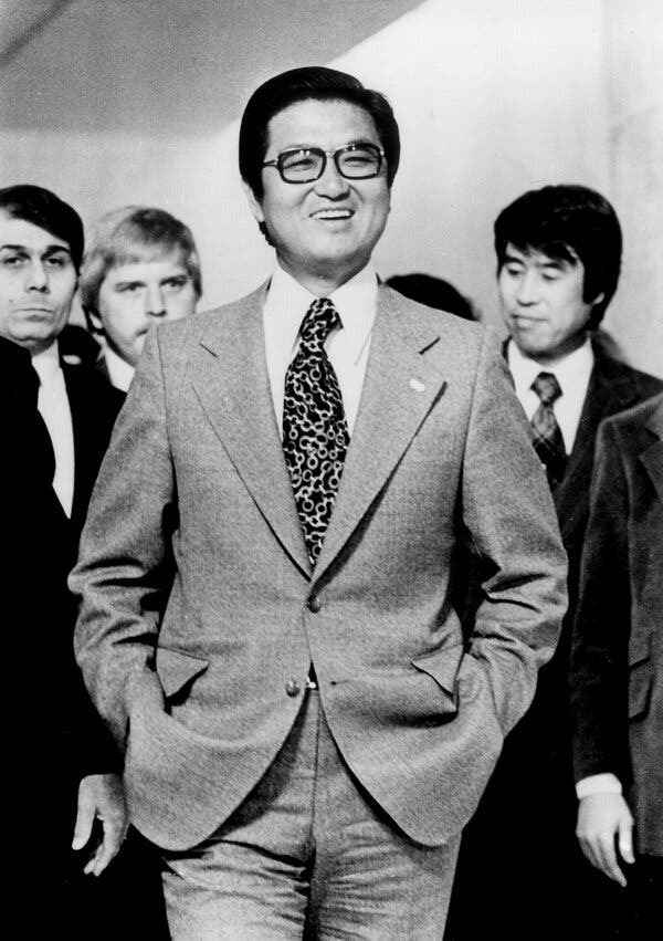 A black-and-white photo of Tongsun Park, a middle-aged man, standing, with several other men behind him. He wears a suit and tie and glasses and has his hands in his pants pockets. He is smiling.