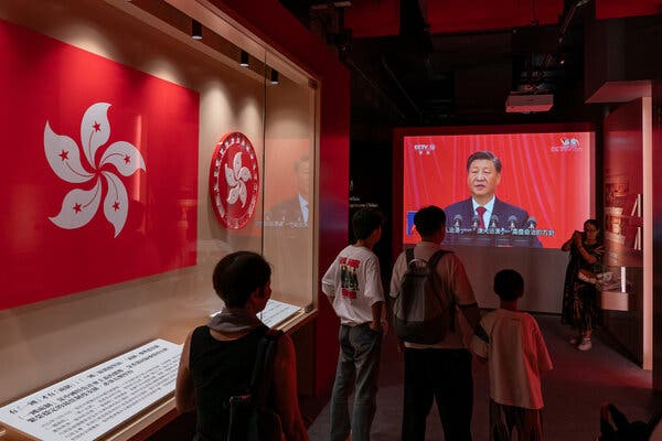 Visitors in a museum look at a screen where Xi Jinping is speaking in front of microphones.