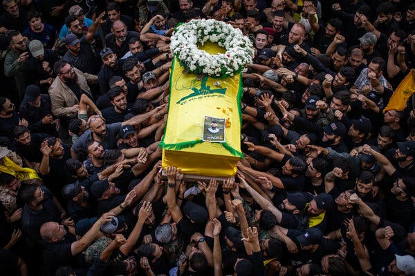 A yellow coffin with a wreath on it being held up by a large group of men.