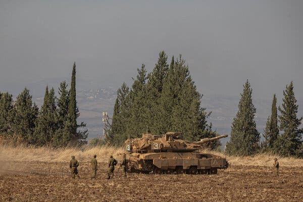 Soldiers next to a tank in a field, with a line of trees behind it and a hillside seen through haze beyond that.