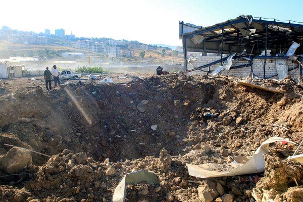 Two people stand at the edge of a large crater. The remains of a building cling to the edge of the crater nearby.