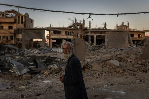 A man in a dark jacket walks through the rubble of destroyed buildings.