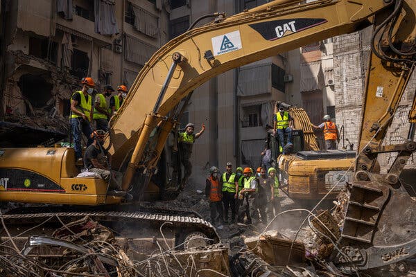 People in high-visibility vests look on as a bulldozer digs through the rubble of a destroyed building.