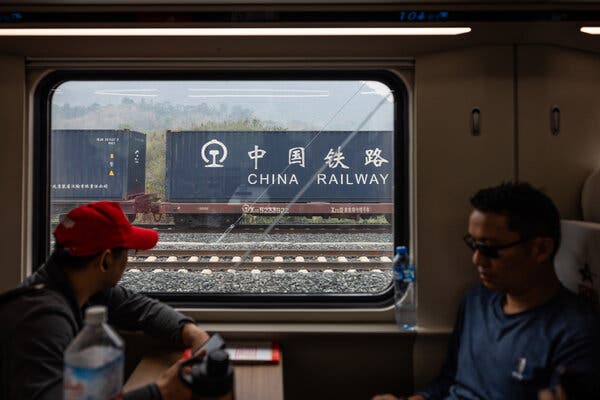 Two train passengers sit facing each other. A China Railway freight train passing by is seen through the window.