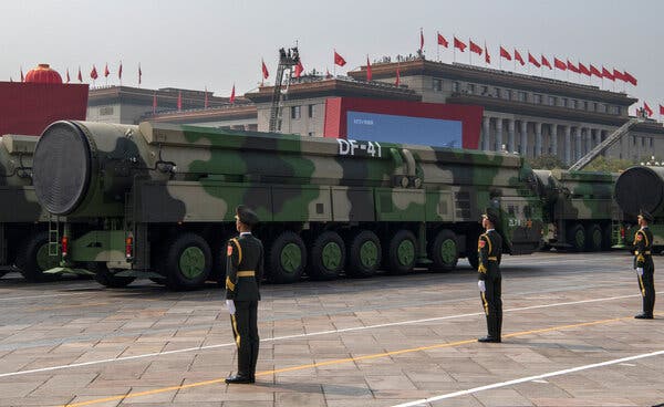 Three men in military uniforms stand at attention in front of a camouflage-colored long-range missile.