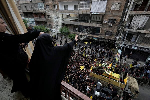 Two people wearing black throw white rice out a window on a crowd of people walking in the street below.