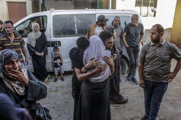 Two people hugging someone, seen from behind, while several others stand nearby in front of a white van. 