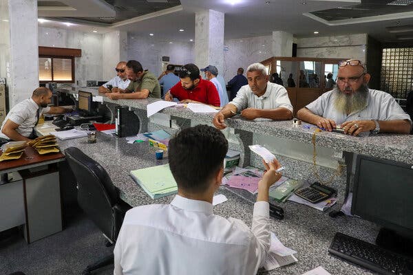 Several people stand along a counter in a bank as two tellers work from behind the counter.