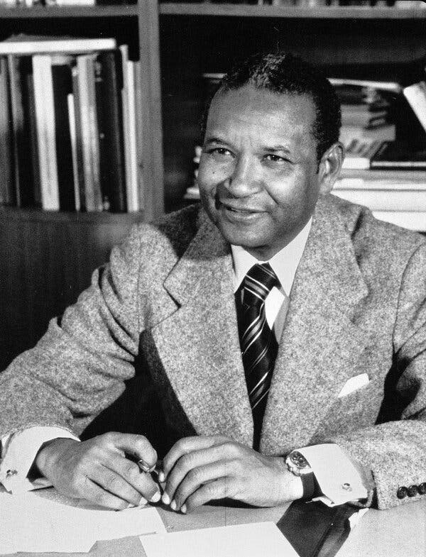 A close-up black and white photo of him sitting at his desk in his office with a half smile. He wears a light-colored wool suit over a white shirt and tie. 