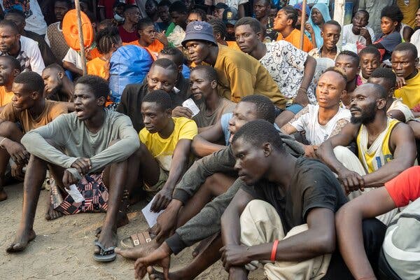 Many casually dressed men sit on the ground, closely packed together.