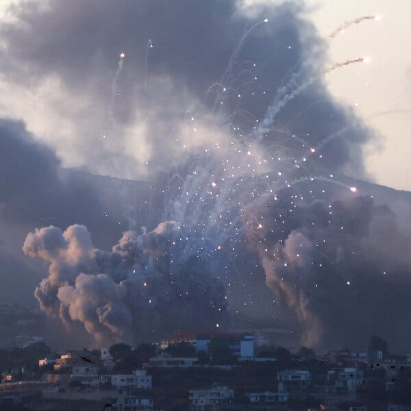 Huge clouds of smoke behind small explosions in the sky over a hill covered with homes.