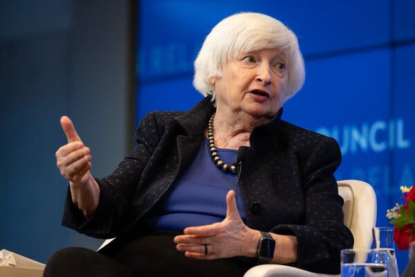 Treasury Secretary Janet L. Yellen, wearing a black jacket and necklace, gestures with her hands while talking on a stage with a blue background.